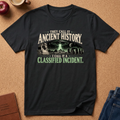 A black graphic t-shirt laid flat on a cork background, featuring a sci-fi design with a UFO, stone circle, and text reading 'THEY CALL IT ANCIENT HISTORY. I CALL IT A CLASSIFIED INCIDENT.