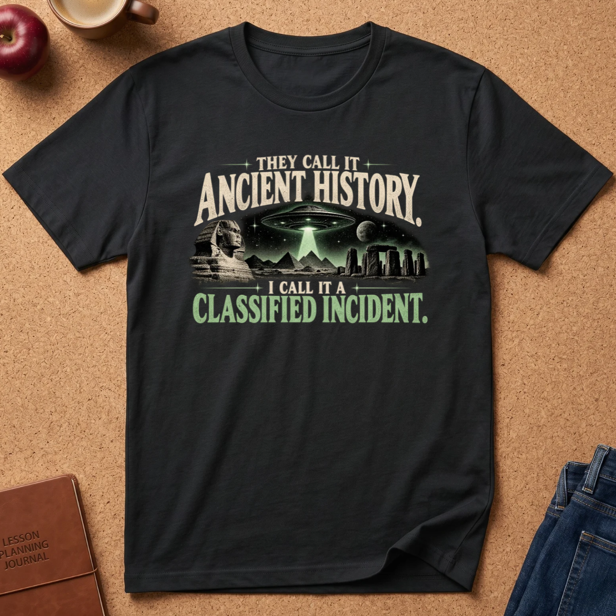 A black graphic t-shirt laid flat on a cork background, featuring a sci-fi design with a UFO, stone circle, and text reading 'THEY CALL IT ANCIENT HISTORY. I CALL IT A CLASSIFIED INCIDENT.