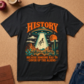 lack  graphic t-shirt laid flat on a cork background, featuring a retro-style alien abduction scene with the text 'HISTORY BECAUSE SOMEONE HAS TO COVER UP THE ALIENS'. The shirt is styled with a brown leather journal, a red apple, a coffee mug, and blue jeans.
