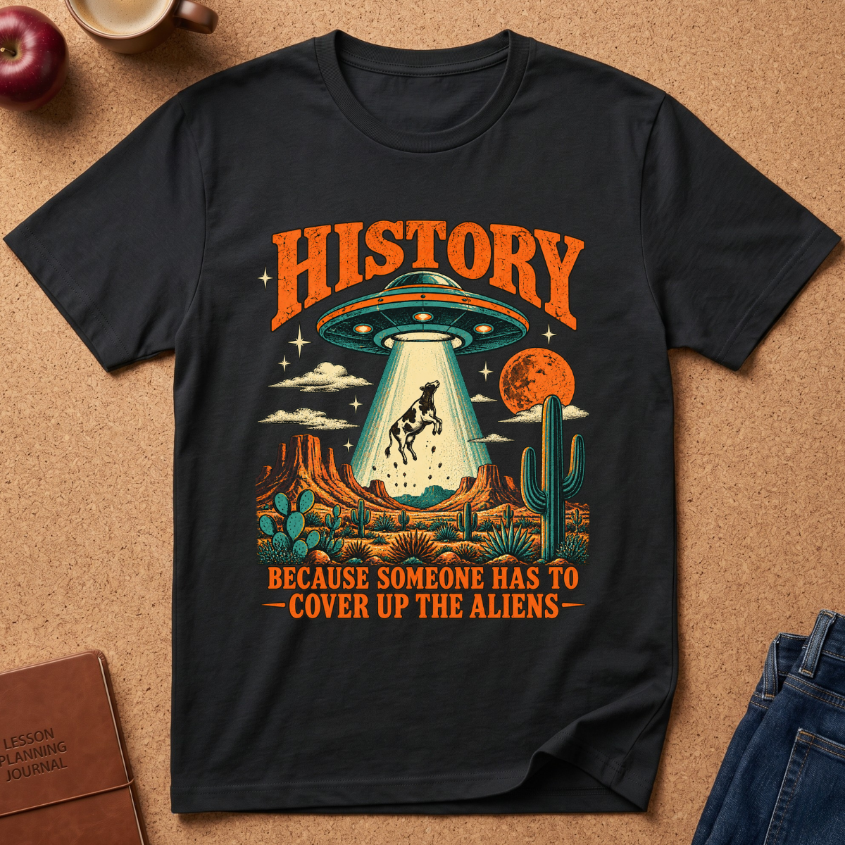 lack  graphic t-shirt laid flat on a cork background, featuring a retro-style alien abduction scene with the text 'HISTORY BECAUSE SOMEONE HAS TO COVER UP THE ALIENS'. The shirt is styled with a brown leather journal, a red apple, a coffee mug, and blue jeans.