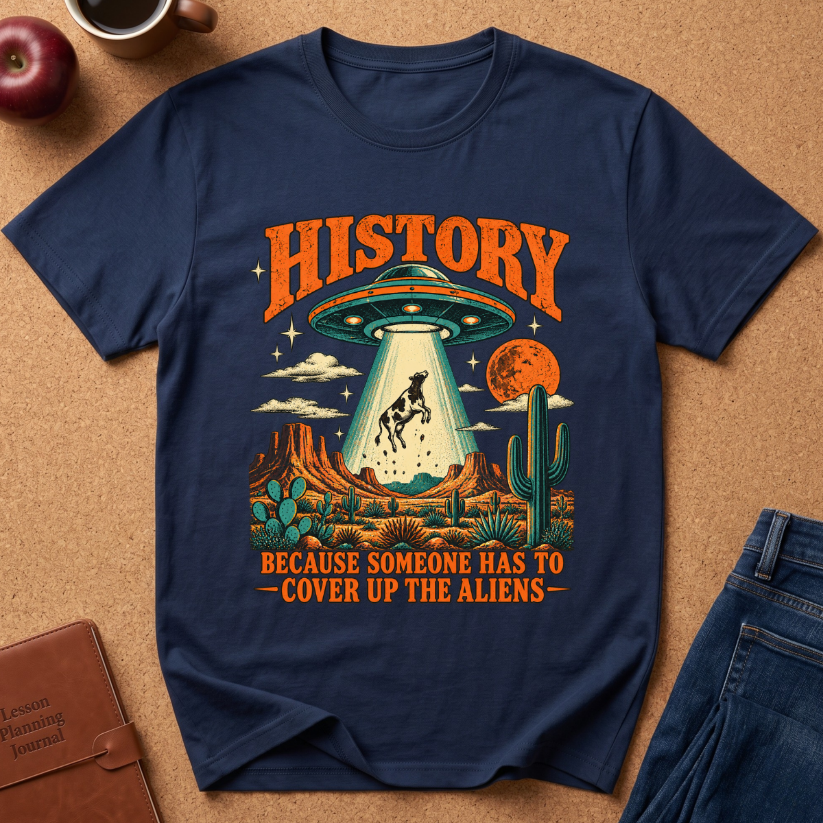 A navy blue graphic t-shirt laid flat on a cork background, featuring a retro-style alien abduction scene with the text 'HISTORY BECAUSE SOMEONE HAS TO COVER UP THE ALIENS'. The shirt is styled with a brown leather journal, a red apple, a coffee mug, and blue jeans.