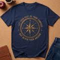 A navy blue short-sleeve t-shirt laid flat on a brown cork background, featuring a large gold compass graphic with the text 'LET YOUR PAST BE YOUR COMPASS BUT NEVER YOUR ANCHOR.' Styled flat-lay props include an apple, a coffee cup, a leather journal, and blue jeans.