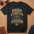 Black  graphic t-shirt laid flat on a cork background, featuring a vintage-style nautical design with the text 'LET THE PAST BE YOUR COMPASS BUT NEVER YOUR ANCHOR' and an anchor and compass motif. Styled flat-lay props include a brown leather journal, a red apple, a coffee mug, and blue jeans.
