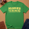 A green short-sleeve T-shirt laid flat on a cork-like surface, with large yellow text reading 'HISTORY TEACHERS' and smaller text below reading 'ALWAYS BRINGING UP THE PAST.' Styled flat-lay props include a brown notebook, an apple, a mug, and blue jeans around the shirt.