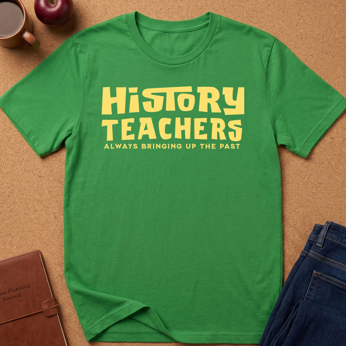 A green short-sleeve T-shirt laid flat on a cork-like surface, with large yellow text reading 'HISTORY TEACHERS' and smaller text below reading 'ALWAYS BRINGING UP THE PAST.' Styled flat-lay props include a brown notebook, an apple, a mug, and blue jeans around the shirt.