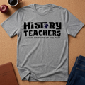 A heather gray short-sleeve T-shirt laid flat on a cork surface, printed with the words 'HISTORY TEACHERS' and 'ALWAYS BRINGING UP THE PAST' in black, with a small globe graphic inside the letter O. Styled flat-lay props include an apple, a coffee cup, a brown journal, and folded blue jeans.