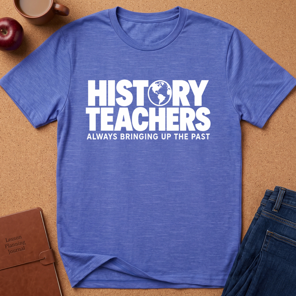 A heathered blue short-sleeve t-shirt with a white graphic text reading 'HISTORY TEACHERS ALWAYS BRINGING UP THE PAST' laid flat on a cork background with a notebook, apple, coffee cup, and jeans as props.