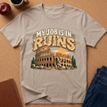A light beige graphic t-shirt laid flat on a cork background, featuring a vintage-style illustration of the Colosseum with the text 'MY JOB IS IN RUINS'. The shirt is styled with a coffee mug, apple, notebook, and blue jeans around it.