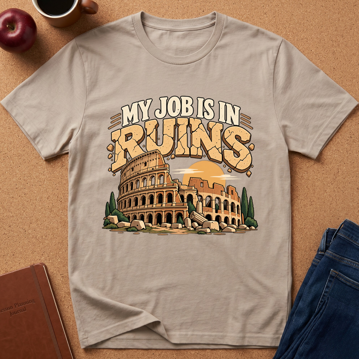 A light beige graphic t-shirt laid flat on a cork background, featuring a vintage-style illustration of the Colosseum with the text 'MY JOB IS IN RUINS'. The shirt is styled with a coffee mug, apple, notebook, and blue jeans around it.