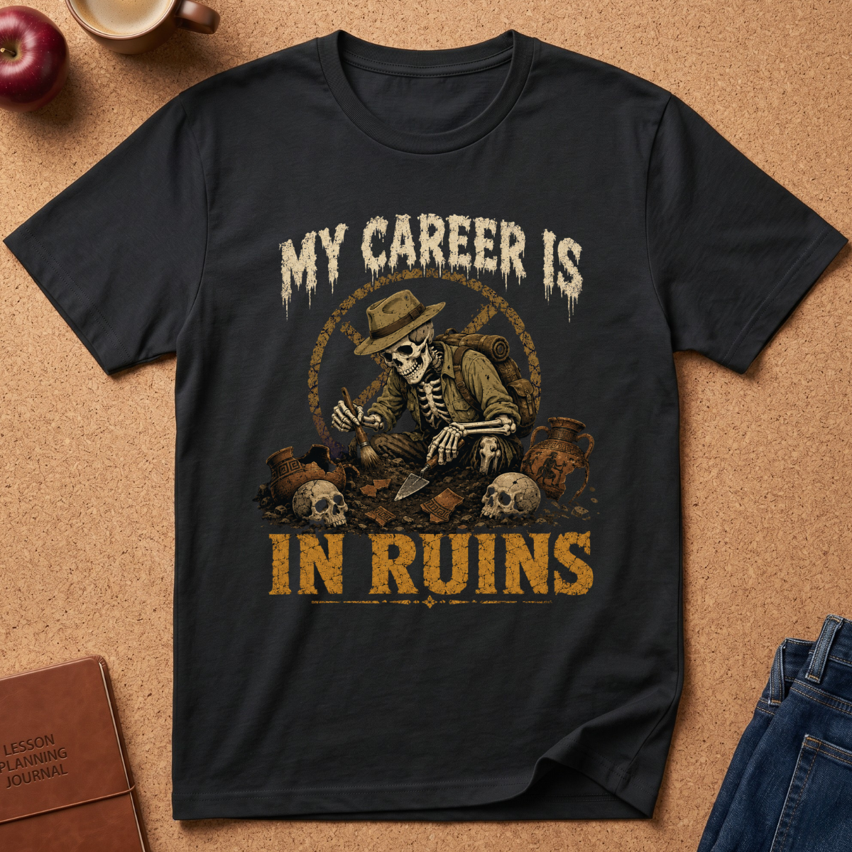 A black graphic t-shirt laid flat on a cork background, featuring a skeleton archaeologist illustration with the text 'MY CAREER IS IN RUINS' in distressed cream and gold lettering. Styled flat-lay props include a brown journal, an apple, a coffee cup, and blue jeans.