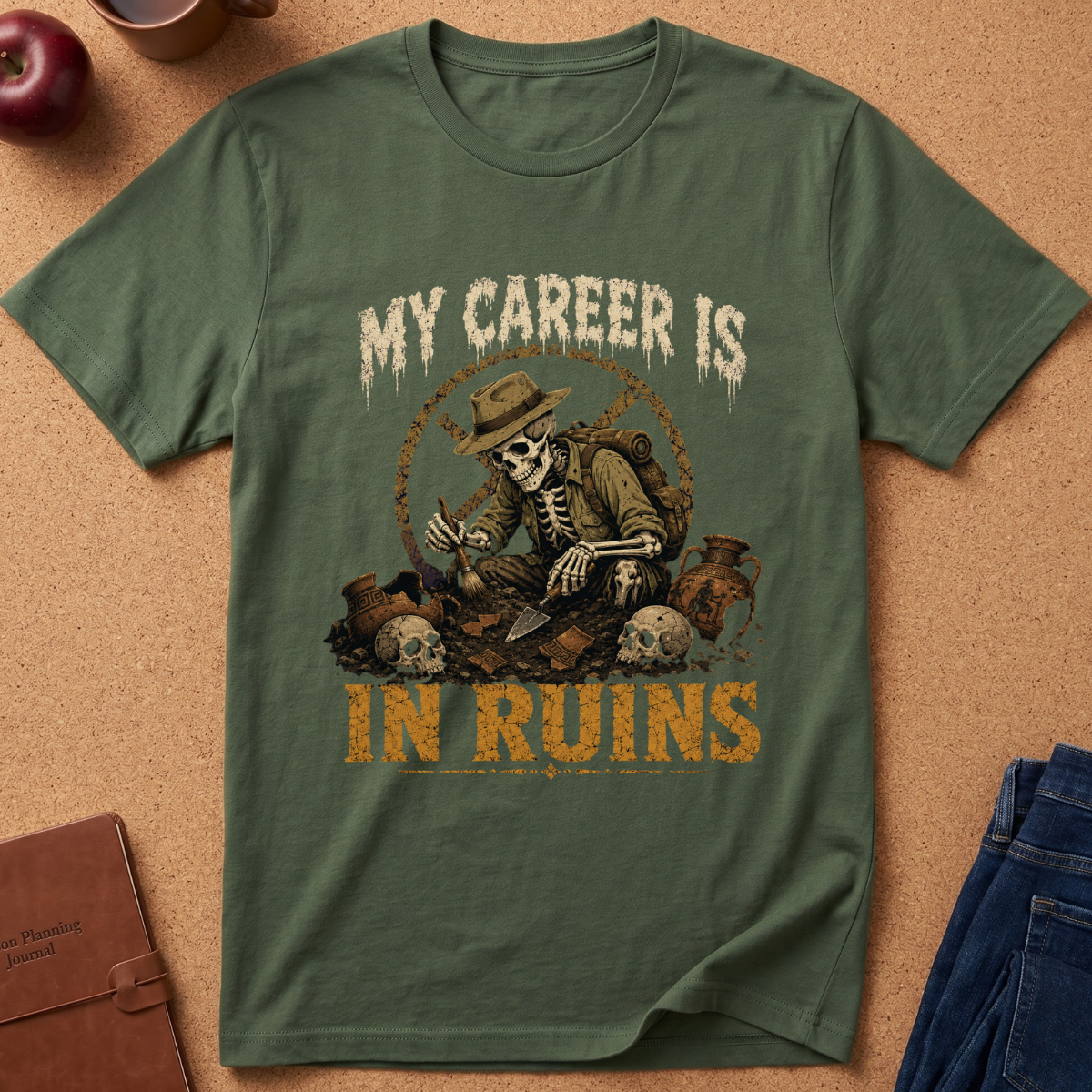 An olive green graphic t-shirt laid flat on a cork background, featuring a skeleton in a hat holding a pickaxe among ruins, with the text 'MY CAREER IS IN RUINS' in distressed cream and orange lettering.