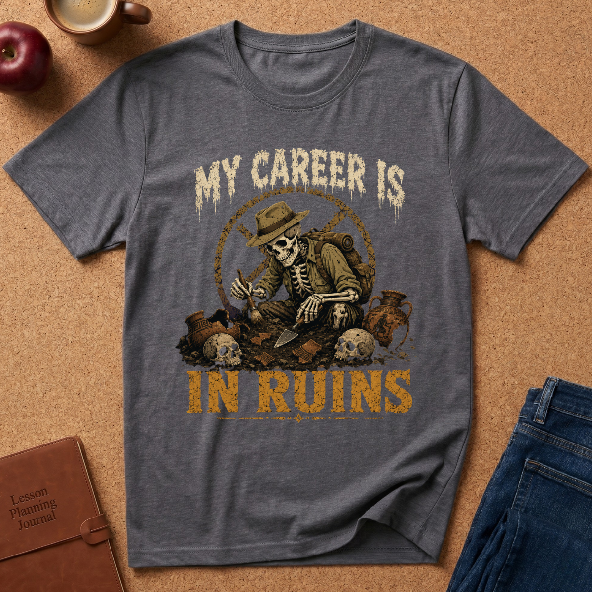 A heather dark gray graphic t-shirt laid flat on a cork background, featuring a skeleton archaeologist illustration with the text 'MY CAREER IS IN RUINS'. The shirt is styled with a leather journal, an apple, and blue jeans.