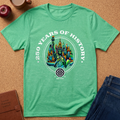 A green graphic t-shirt laid flat on a cork background, featuring a colorful Statue of Liberty and New York City skyline design with the text '250 YEARS OF HISTORY'. Props like an apple, coffee cup, notebook, and jeans are visible around the shirt.