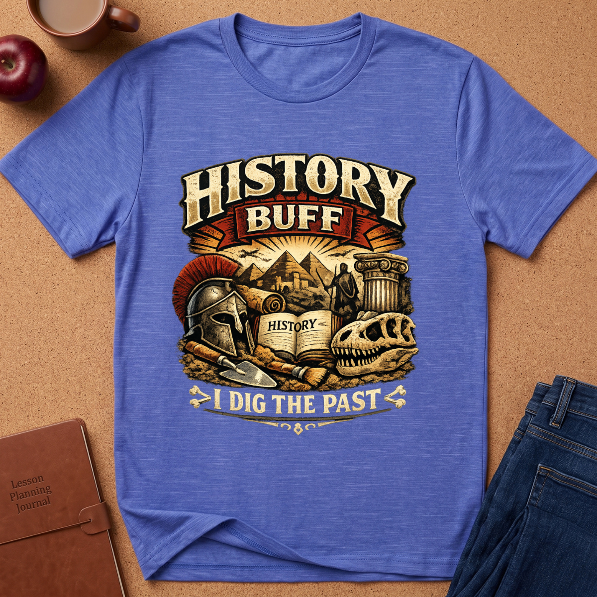 A heather blue graphic t-shirt laid flat on a cork background, surrounded by lifestyle props including a brown leather journal, a red apple, a coffee mug, and blue jeans. The t-shirt features a detailed graphic with the text 'HISTORY BUFF I DIG THE PAST' and illustrations of a gladiator helmet, an open book labeled 'HISTORY', a column, and a skull.