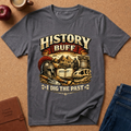 A heather dark gray graphic t-shirt laid flat on a cork background, surrounded by lifestyle props including an apple, a coffee cup, a leather journal, and blue jeans. The t-shirt features a vintage-style graphic with the text 'HISTORY BUFF I DIG THE PAST' and illustrations of a gladiator helmet, scroll, and ancient ruins.