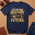 A navy blue graphic t-shirt laid flat on a cork background, featuring the text 'STUDYING HISTORY IS KNOWING THE FUTURE' in gold and cream lettering with an illustration of an old book and an hourglass. Styled flat-lay props include a brown leather journal, a red apple, a coffee cup, and blue jeans.