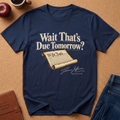 A navy blue graphic t-shirt laid flat on a cork background, featuring the text 'Wait That's Due Tomorrow?' and a scroll graphic with 'We the People' and 'Thomas Jefferson July 4th 1776'.