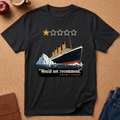 A black graphic t-shirt laid flat on a cork background, featuring an illustration of the Titanic with a iceberg and the text 'Would not recommend' with 'Anonymous Passenger' underneath. Styled flat-lay props include an apple, a coffee mug, a journal, and jeans.