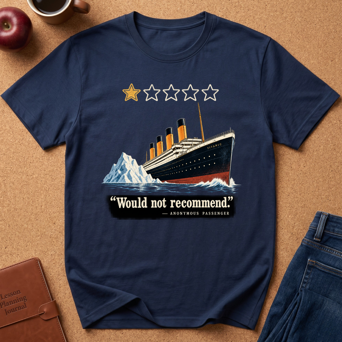 A navy blue graphic t-shirt laid flat on a brown cork background, featuring an illustration of the Titanic with a iceberg and the text 'Would not recommend' with a five-star rating graphic. Styled flat-lay props include a brown leather journal, a red apple, a coffee cup, and blue jeans.