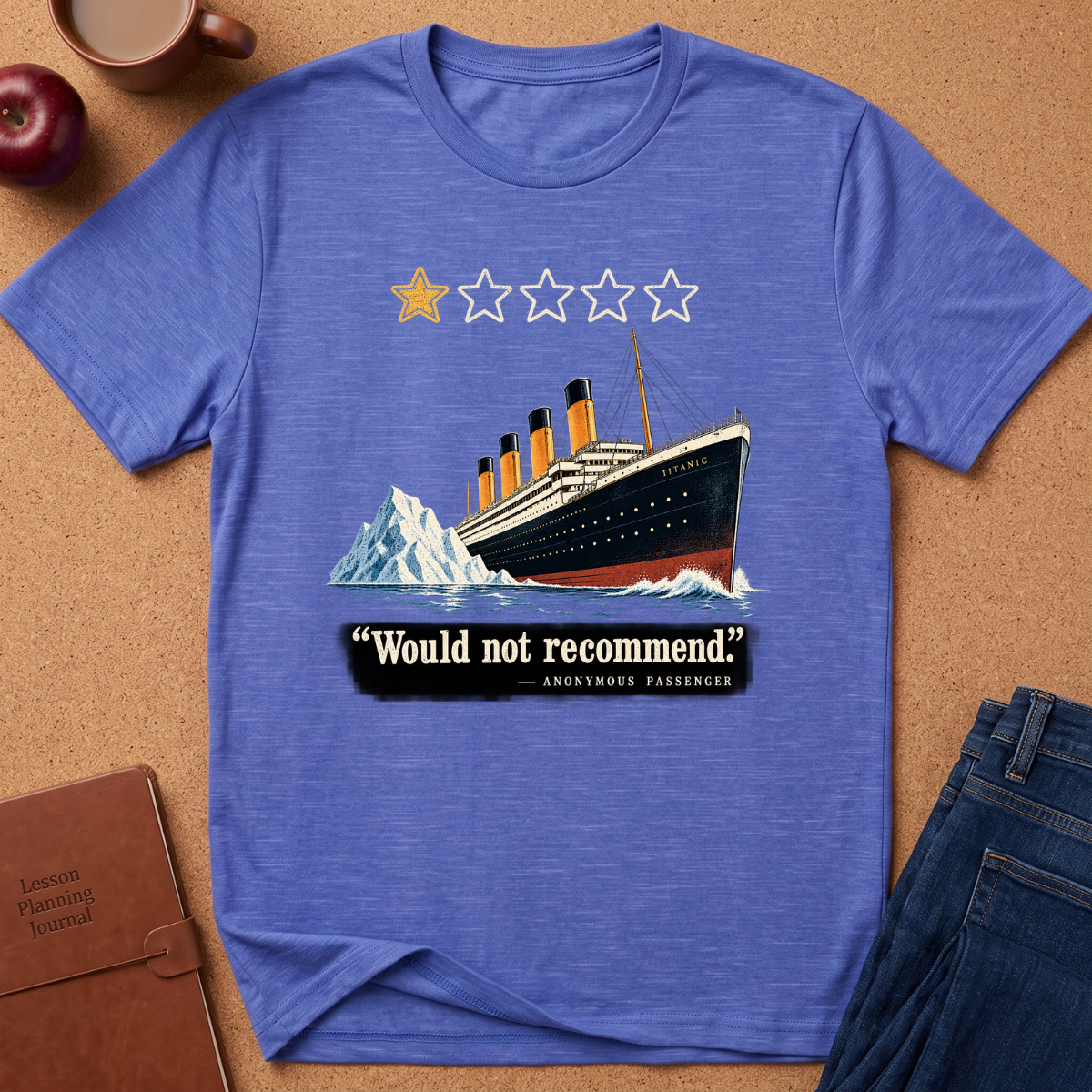 A heathered blue short-sleeve t-shirt laid flat on a cork background, featuring a Titanic graphic with a one-star rating and the quote 'Would not recommend' from Anonymous Passenger. Styled flat-lay props include an apple, coffee cup, notebook, and jeans.