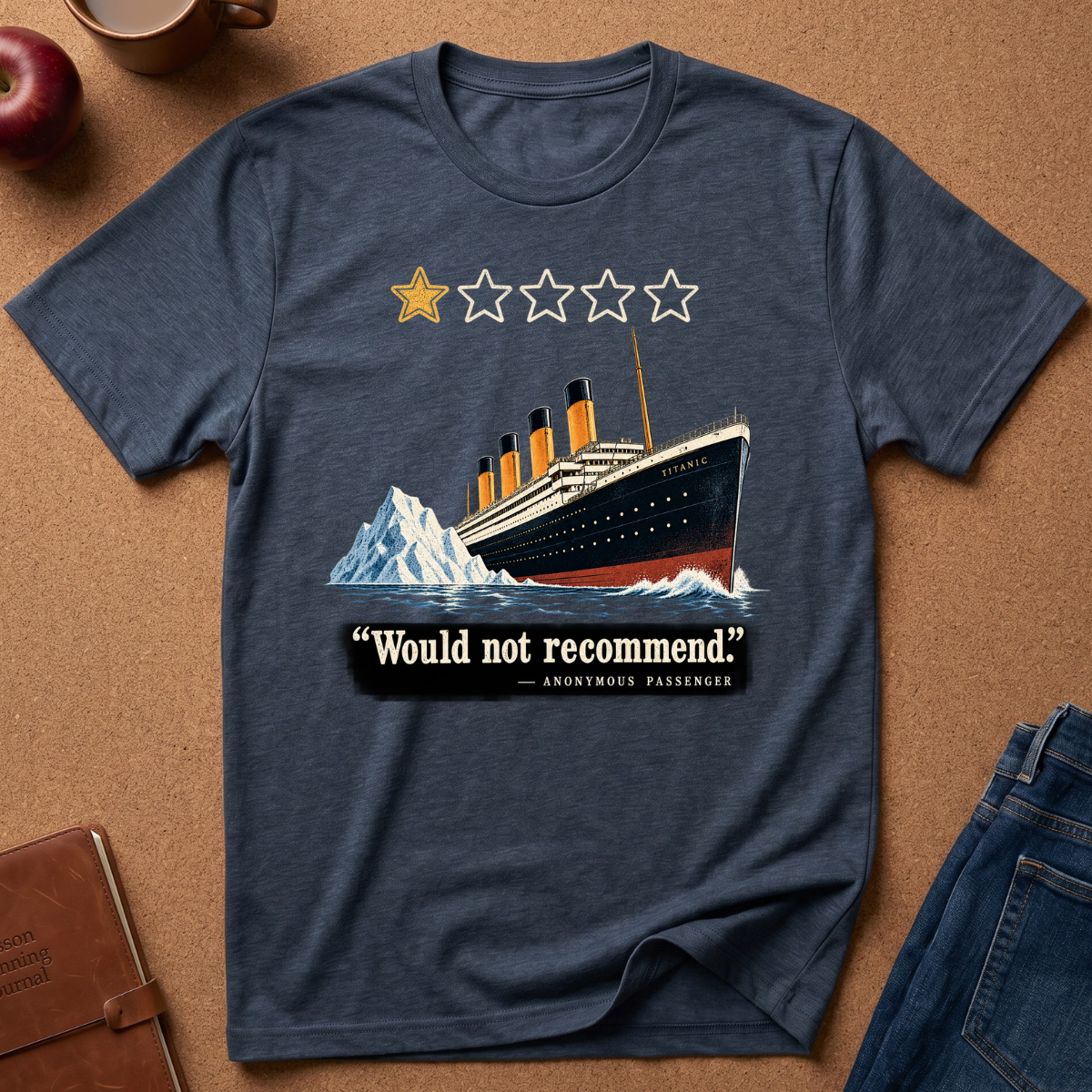 A heather navy blue graphic t-shirt laid flat on a cork background with lifestyle props like an apple, a coffee mug, a notebook, and jeans. The graphic features a Titanic ship, a mountain, a 1-star review icon, and the quote 'Would not recommend' - Anonymous Passenger.