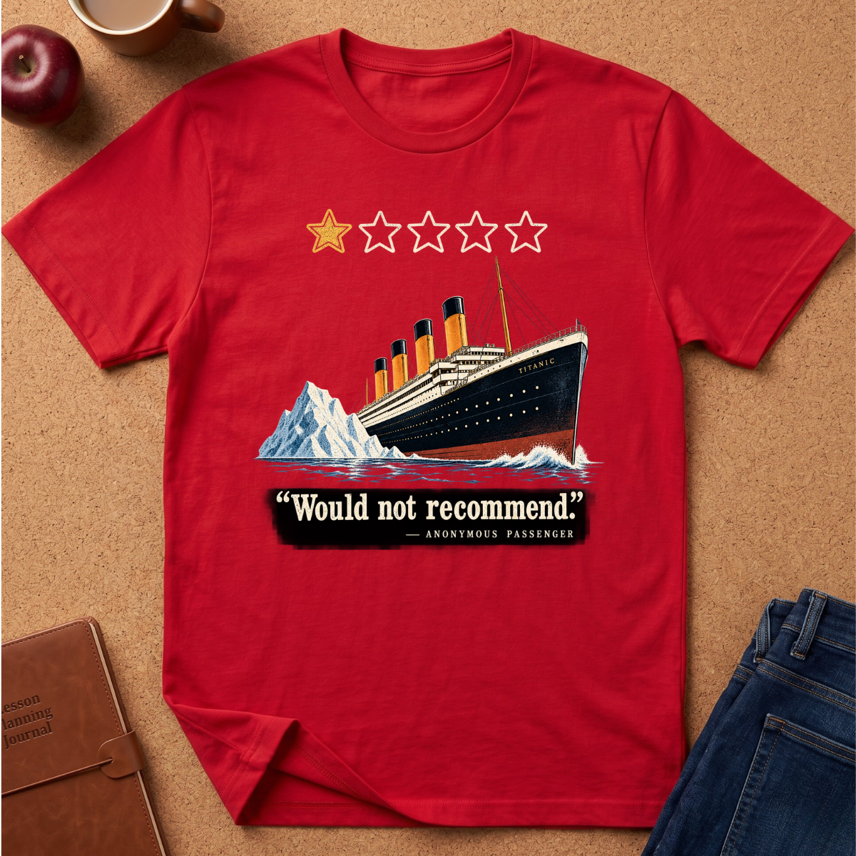 A red short-sleeve graphic T-shirt laid flat on a cork surface, featuring an illustration of the Titanic with a iceberg, four black-and-red smokestacks, a yellow star, three white stars, and the text “Would not recommend” with “Anonymous Passenger” underneath. Partial props including an apple, a mug, a notebook, and jeans are visible around the shirt.