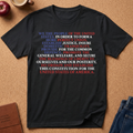 A black graphic t-shirt laid flat on a cork background, featuring a multi-colored patriotic text design in blue, purple, and red. The shirt is styled with a brown leather journal, an apple, a coffee mug, and blue jeans.