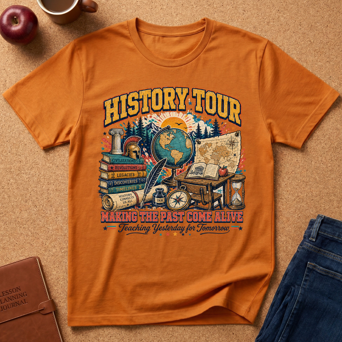 An orange graphic t-shirt laid flat on a brown cork surface, surrounded by lifestyle props including a brown leather journal, a red apple, a coffee mug, and blue jeans. The t-shirt features a colorful vintage-style 'HISTORY TOUR' educational graphic with globes, maps, books, and compasses.