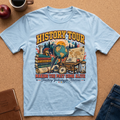 A Light Blue graphic t-shirt laid flat on a brown cork surface, surrounded by lifestyle props including a brown leather journal, a red apple, a coffee mug, and blue jeans. The t-shirt features a colorful vintage-style 'HISTORY TOUR' educational graphic with globes, maps, books, and compasses.