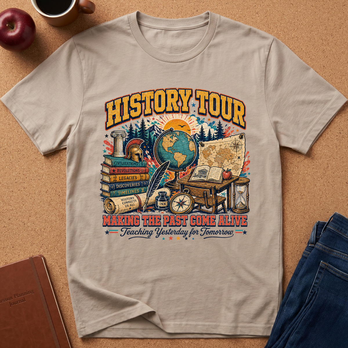 A Sand colored graphic t-shirt laid flat on a brown cork surface, surrounded by lifestyle props including a brown leather journal, a red apple, a coffee mug, and blue jeans. The t-shirt features a colorful vintage-style 'HISTORY TOUR' educational graphic with globes, maps, books, and compasses.