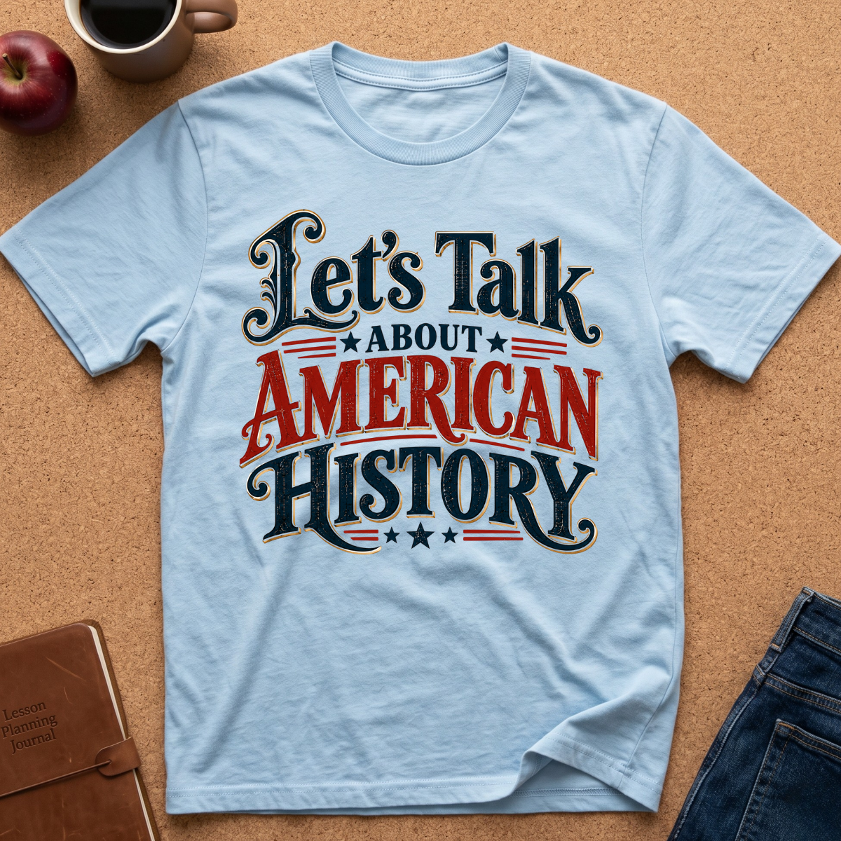 A light blue graphic t-shirt laid flat on a cork background, featuring the text 'Let's Talk ABOUT AMERICAN HISTORY' in black and red lettering. Styled flat-lay props include a coffee mug, an apple, a notebook, and jeans.