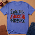 A heather blue short-sleeve t-shirt with a graphic reading 'Let's Talk ABOUT AMERICAN HISTORY' in dark teal and red lettering, styled in a flat-lay on a cork background with an apple, coffee mug, notebook, and jeans nearby.