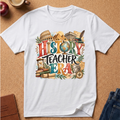 A white short-sleeve graphic T-shirt laid flat on a cork surface, featuring a colorful educational design with the words “HISTORY TEACHER ERA” and illustrations of landmarks, books, scrolls, and a globe.