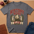 A gray short-sleeve graphic T-shirt laid flat on a cork surface, featuring the words “HISTORY NERD CLUB” in red and cream with a globe, books, laurel wreath, and columns. Surrounding props include an apple, coffee cup, notebook, and jeans.