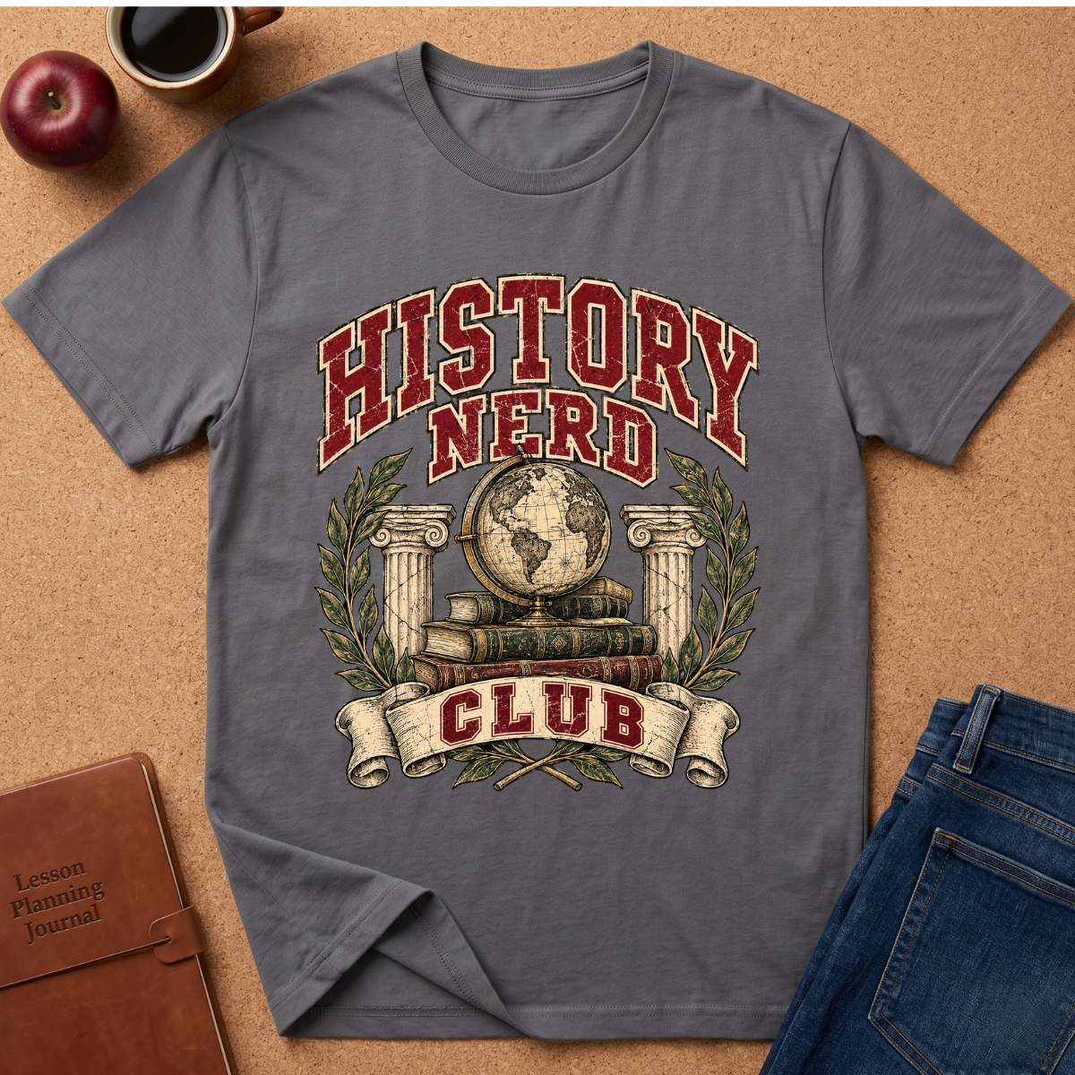 A gray short-sleeve graphic T-shirt laid flat on a cork surface, featuring the words “HISTORY NERD CLUB” in red and cream with a globe, books, laurel wreath, and columns. Surrounding props include an apple, coffee cup, notebook, and jeans.