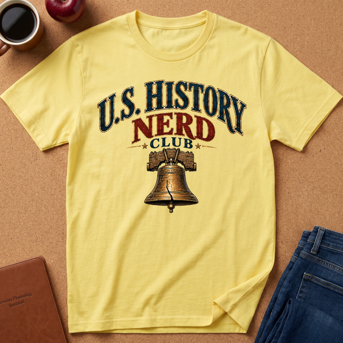 A pale yellow graphic T-shirt laid flat on a cork-like surface, printed with the words 'U.S. History Nerd Club" With an image of the liberty bell.