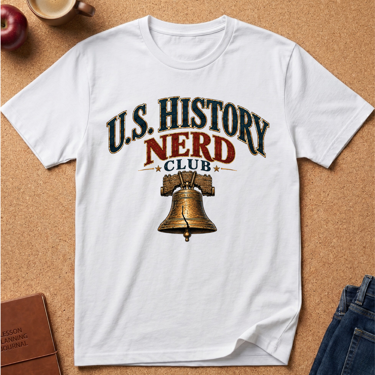 A white short-sleeve graphic T-shirt laid flat on a cork surface, featuring the text 'U.S. History Nerd Club" With an image of the liberty bell.