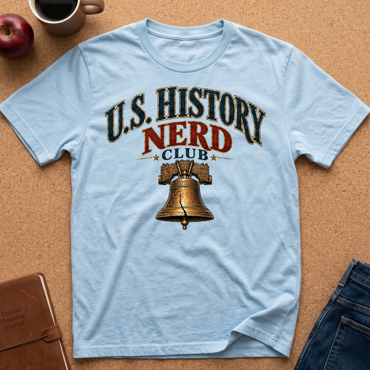 A light blue short-sleeve t-shirt laid flat on a cork background, featuring a graphic print that reads 'U.S. History Nerd Club" With an image of the liberty bell.