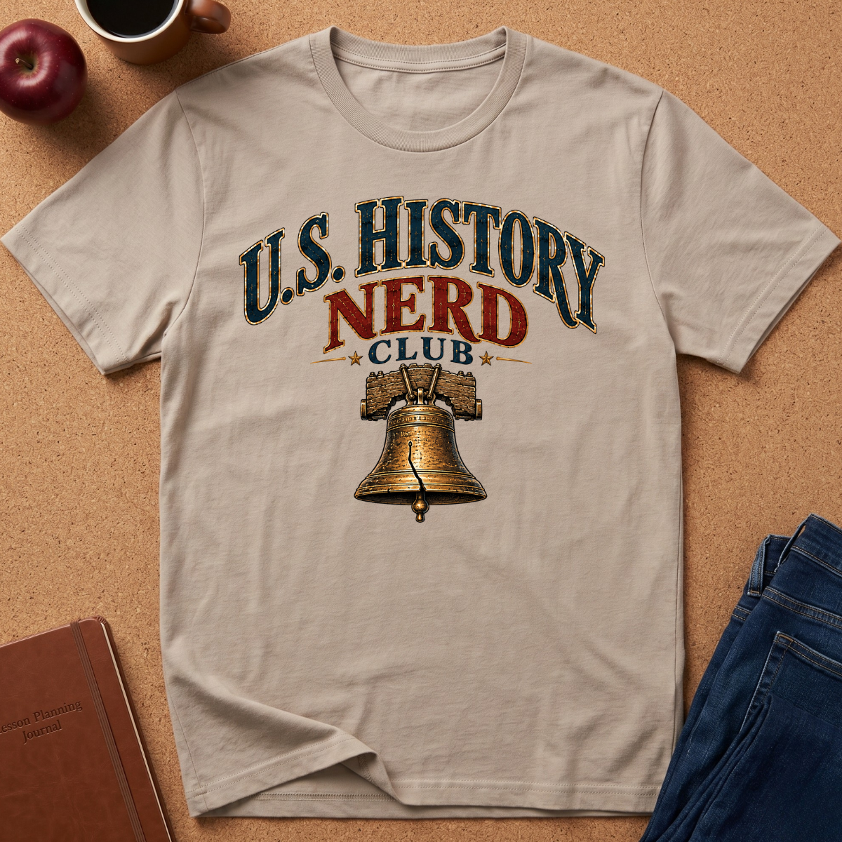 A beige graphic t-shirt laid flat on a cork background, featuring the text 'U.S. History Nerd Club" With an image of the liberty bell.