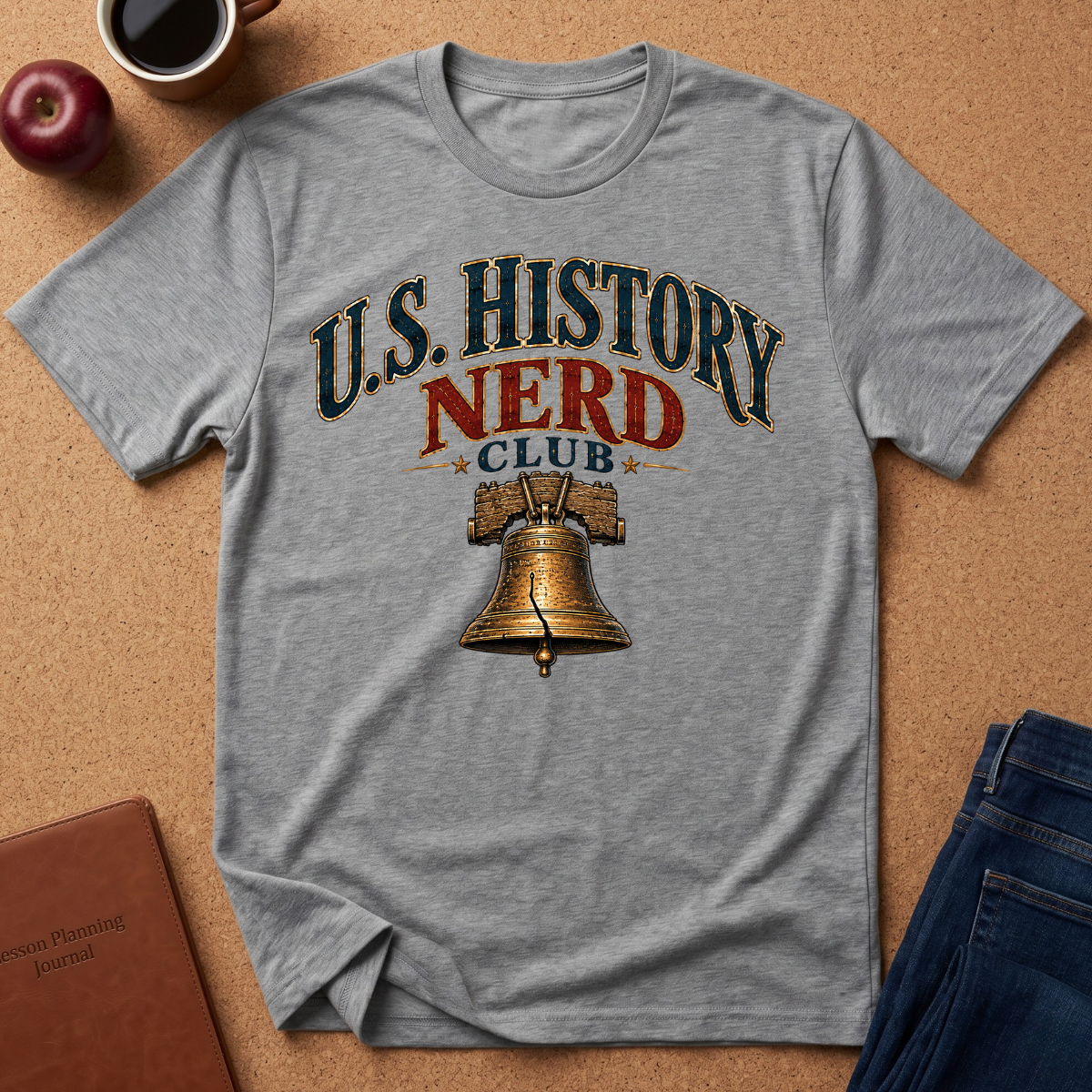 A heather gray graphic t-shirt laid flat on a cork background, featuring a distressed print that reads 'U.S. History Nerd Club" With an image of the liberty bell.
