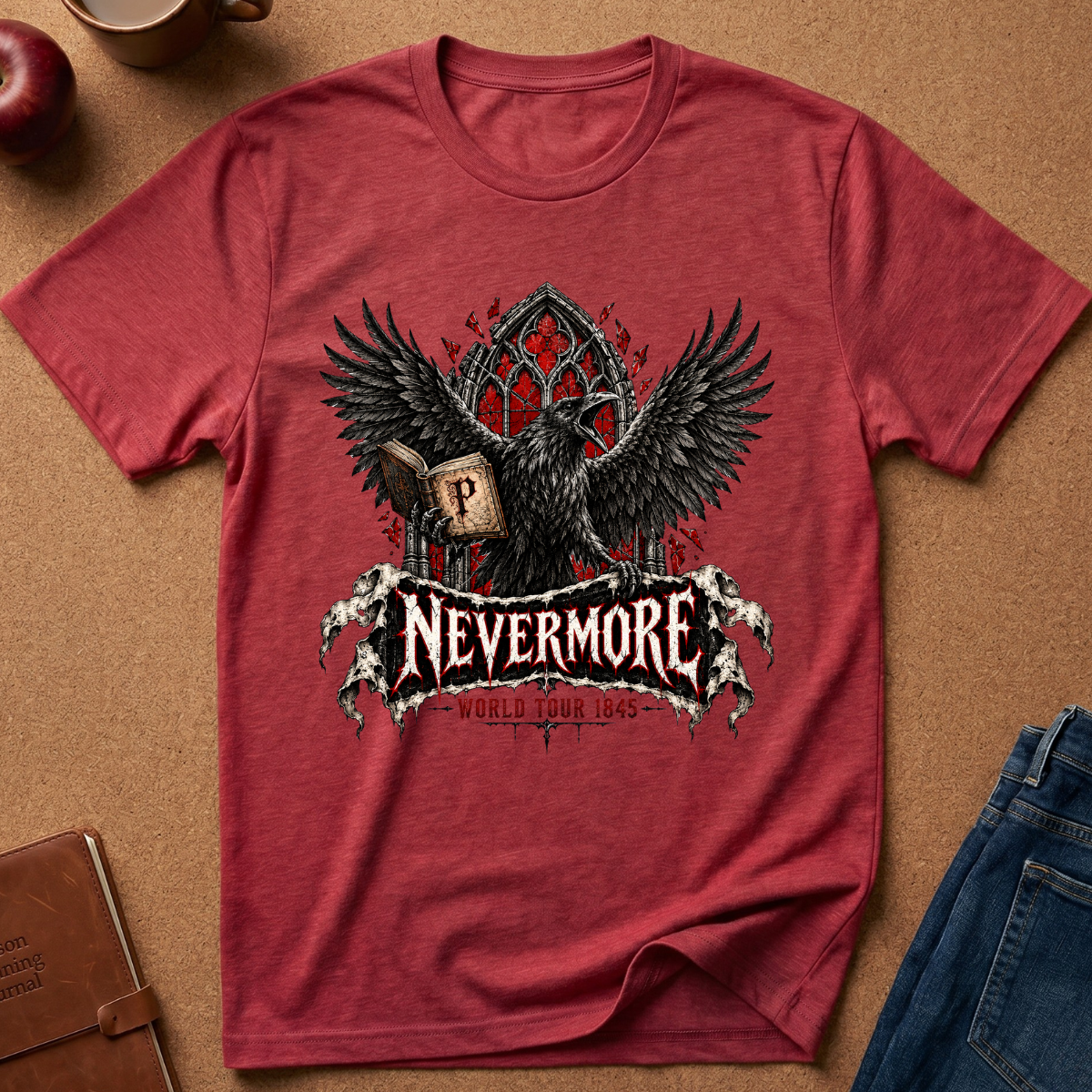 A heathered red short-sleeve t-shirt with a graphic of a raven, a book, and the word 'Nevermore' in a gothic font, displayed flat on a brown textured surface with jeans, a notebook, and an apple partially visible.