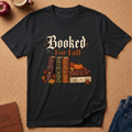 A black short-sleeve graphic T-shirt laid flat on a cork-like surface, featuring the phrase 'Booked For Fall' above a stack of vintage books, pumpkins, and autumn leaves. Partial props around the shirt include a mug, apple, journal, and jeans.