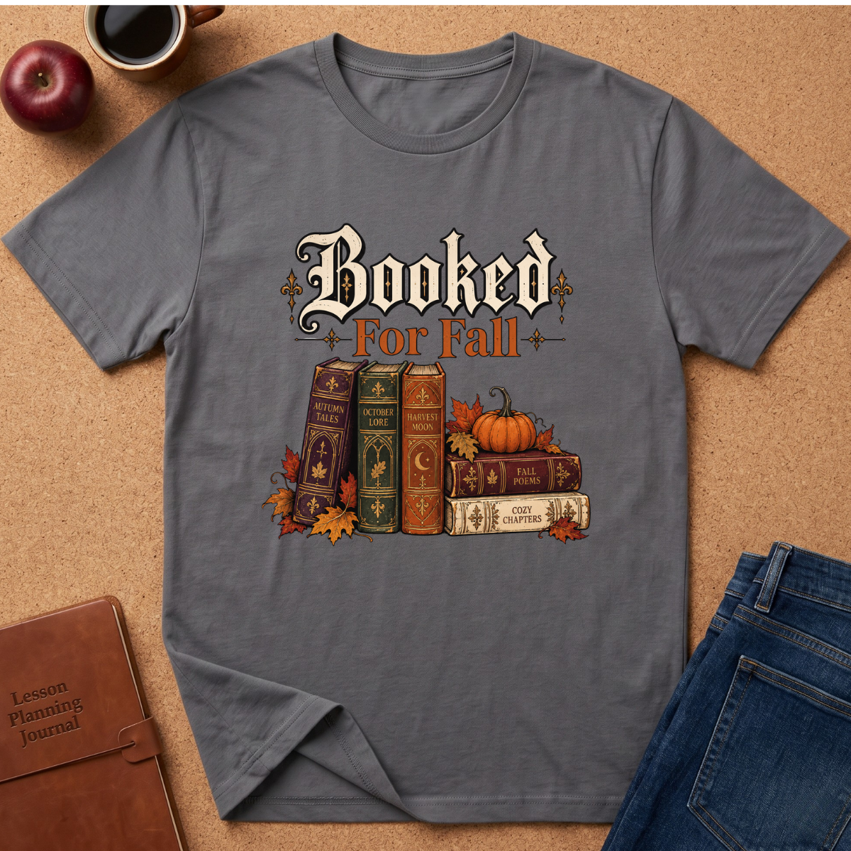 A gray short-sleeve t-shirt laid flat on a cork background, featuring a 'Booked For Fall' graphic with vintage books, pumpkins, and autumn leaves. Styled flat-lay props include a coffee cup, apple, leather journal, and blue jeans.