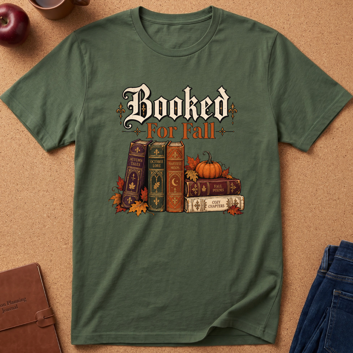An olive green short-sleeve t-shirt laid flat on a cork background, featuring a graphic print that reads 'Booked For Fall' above a stack of vintage books, pumpkins, and autumn leaves.