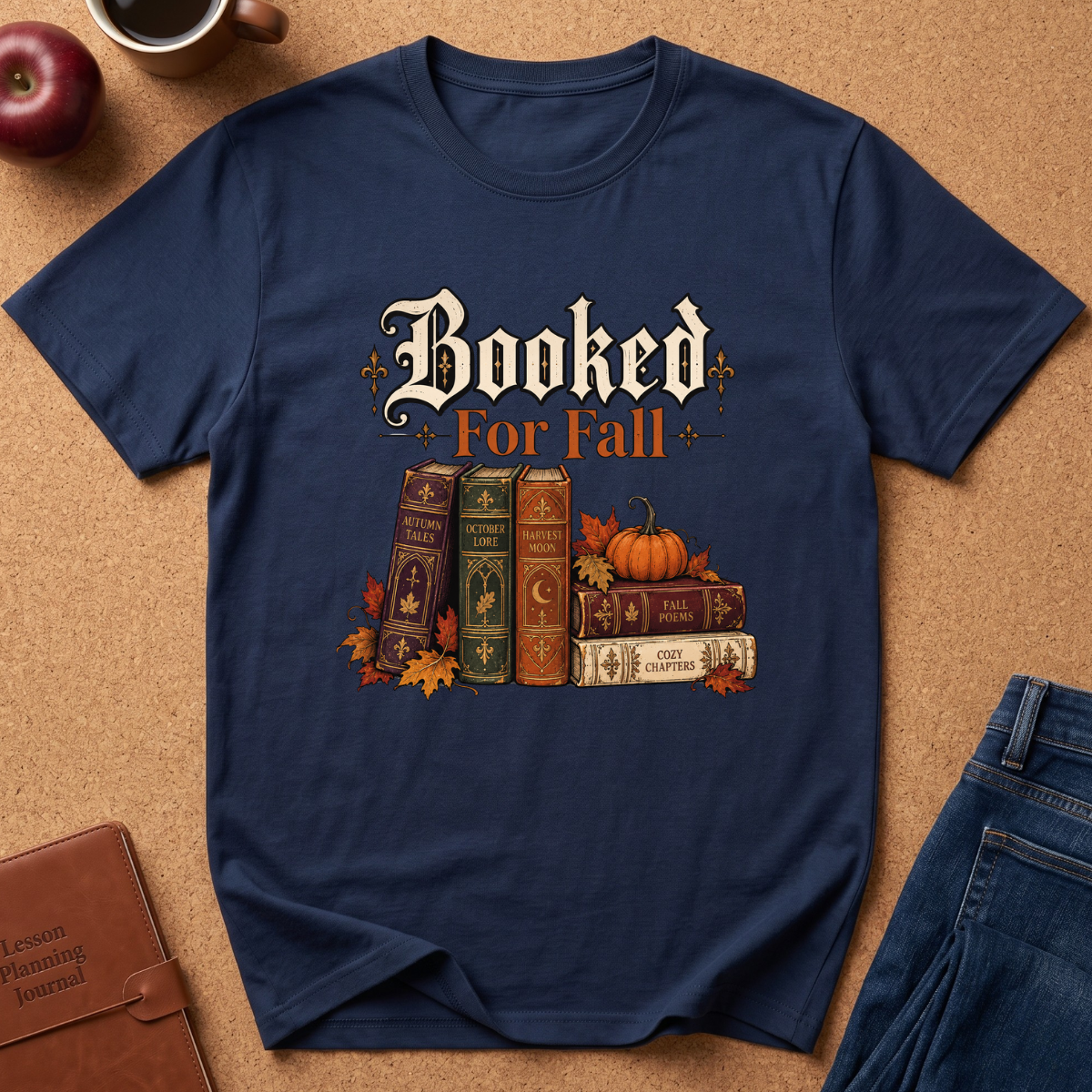 A navy blue graphic t-shirt laid flat on a brown cork background, featuring the text 'Booked For Fall' above an illustration of vintage books, pumpkins, and autumn leaves. Partial props around the shirt include an apple, a coffee cup, a journal, and jeans.