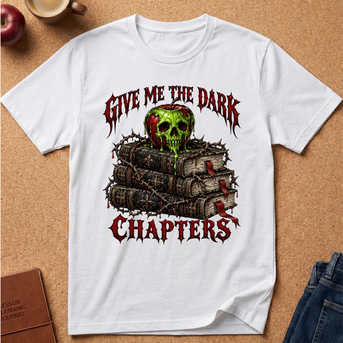 A white short-sleeve graphic T-shirt laid flat on a cork background, featuring the phrase 'GIVE ME THE DARK CHAPTERS' in red gothic lettering around an illustration of stacked books topped with a green-and-red skull and barbed wire.