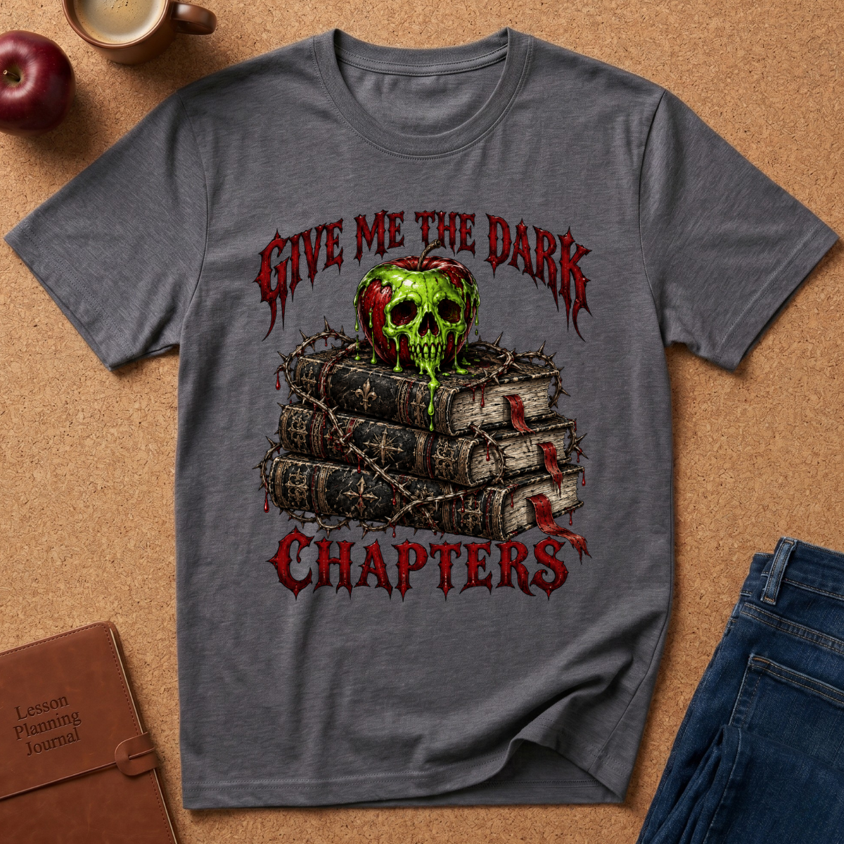 A heather gray graphic t-shirt laid flat on a cork background, featuring a design of a green skull on stacked books with red gothic text reading 'GIVE ME THE DARK CHAPTERS'.