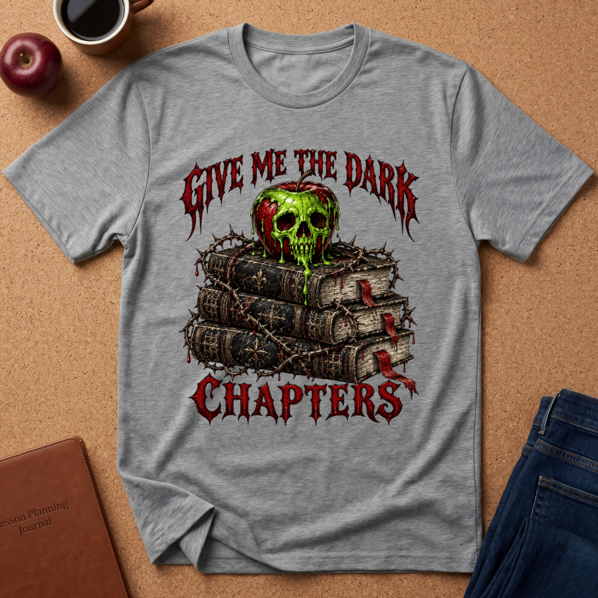 A heather gray graphic t-shirt laid flat on a cork background, featuring a design of a green skull on top of stacked books with barbed wire and the text 'GIVE ME THE DARK CHAPTERS' in red. Lifestyle props like a coffee cup, apple, journal, and jeans are visible around the shirt.