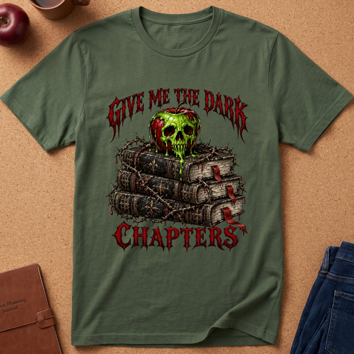 An olive green graphic t-shirt laid flat on a cork background, featuring a design of a green skull on top of stacked books with the gothic text 'GIVE ME THE DARK CHAPTERS'.