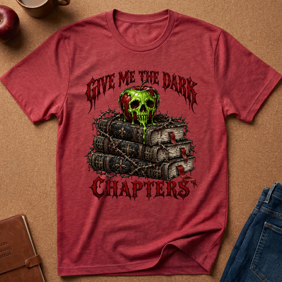 A heathered red graphic t-shirt laid flat on a brown cork background, featuring a green skull on top of dark books with the gothic text 'GIVE ME THE DARK CHAPTERS'.